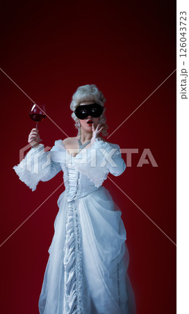 Whimsical duchess. Young woman in rococo dress with glass of red wine and black mask expressing surprise. 126043723