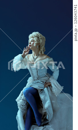 Noble whisper. Young woman in rococo dress with elaborate hairstyle softly gesturing with intrigue against blue background. Noble whisper. Young woman in rococo dress with elaborate hairstyle softly gesturing with intrigue against blue background. 126043741