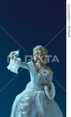 Noble lady in vintage gown and white wig taking selfie with phone and mirror against blue background. Social media post, blogger Noble lady in vintage gown and white wig taking selfie with phone and mirror against blue background. Social media post, blogger 126043742