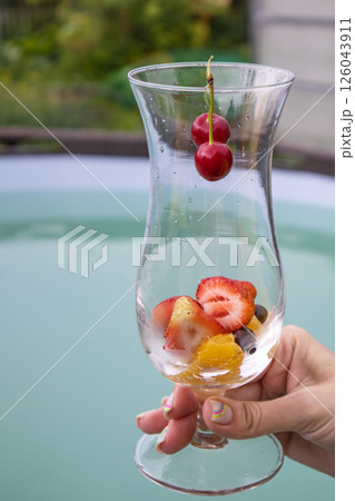 A summer cocktail with strawberries, cherries and foamy champagne in a woman's hand, against a blue pool backdrop. Party concept, vertical 126043911