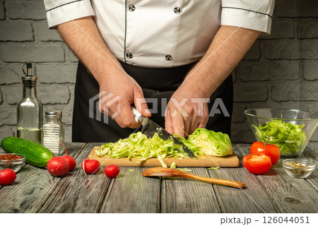 A chef skillfully slices cabbage on a wooden cutting board, surrounded by fresh vegetables like tomatoes and cucumbers, in a stylish kitchen setting A chef skillfully slices cabbage on a wooden cutting board, surrounded by fresh vegetables like tomatoes and cucumbers, in a stylish kitchen setting 126044051