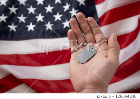 Mans hand holds military soldiers tag against backdrop of American flag. National pride and military tribute with focus on tag. Memorial Day. 126044830