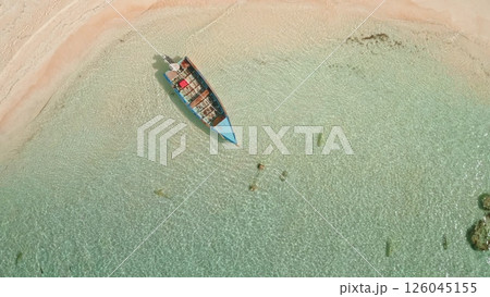 Old wooden colorful boat gently rests on clear turquoise water waves near tropical wild island sandy beach. Coral reef sharks swim around. Exotic travel getaway paradise. Aerial top down zoom out 126045155