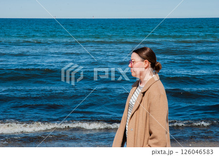 Female adult with glasses strolling on a coastal beachfront, serenity of ocean waves in contrast to clear blue sky for a peaceful day. Digital nomad lifestyle. Remote work 126046583