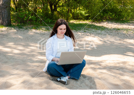 Caucasian young adult female in casual attire focuses on laptop while seated on a sunny sandy beach under the shade of lush green trees. Digital nomad lifestyle. Remote work Caucasian young adult female in casual attire focuses on laptop while seated on a sunny sandy beach under the shade of lush green trees. Digital nomad lifestyle. Remote work 126046655