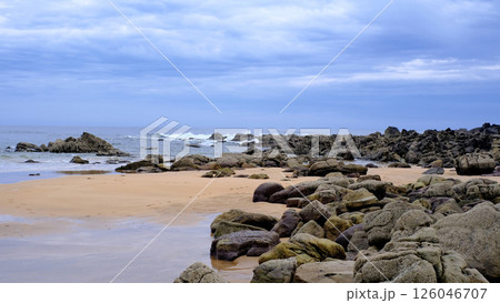 Serene coastal landscape with a wide sandy beach scattered with rugged rocks under a moody cloudy sky 126046707