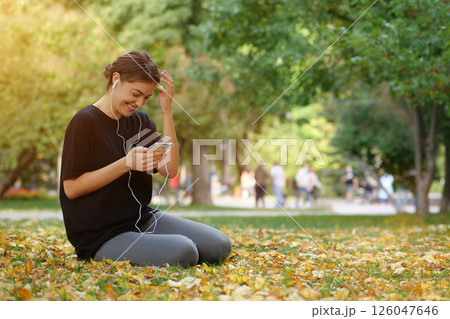 Young beautiful girl sits in park, uses smartphone, listens to music and laughs Young beautiful girl sits in park, uses smartphone, listens to music and laughs 126047646