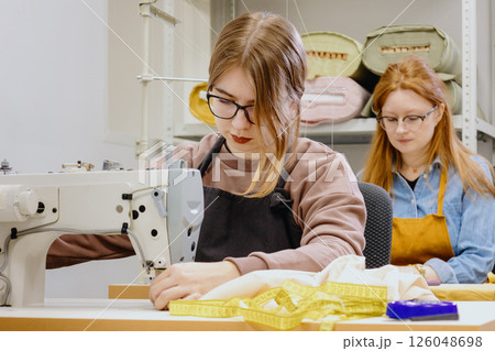Two female seamstresses in the workshop at the factory are working at tables on sewing machines. Two female seamstresses in the workshop at the factory are working at tables on sewing machines. 126048698