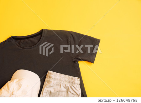 Stylish dark and light grey children's T-shirt with trousers and black cap. Fashion kids outfit for summer, autumn. Total grey look on yellow background. Flat lay, top view Stylish dark and light grey children's T-shirt with trousers and black cap. Fashion kids outfit for summer, autumn. Total grey look on yellow background. Flat lay, top view 126048768