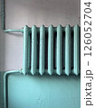 An old central heating radiator in the entrance of an apartment building. The radiator is blue-green in cracked paint against a plaster wall. Heating of buildings during the cold winter season 126052704