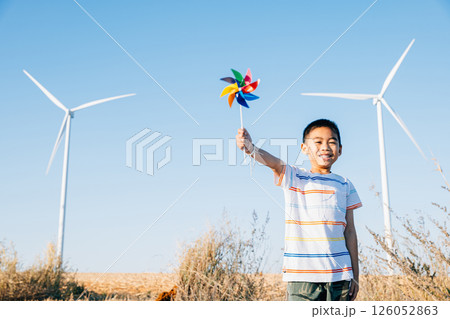 Boy's playful engagement near wind turbines holding a pinwheel toy represents fun of wind energy. Showcases clean electricity innovation in a countryside windmill landscape against a clear blue sky. 126052863
