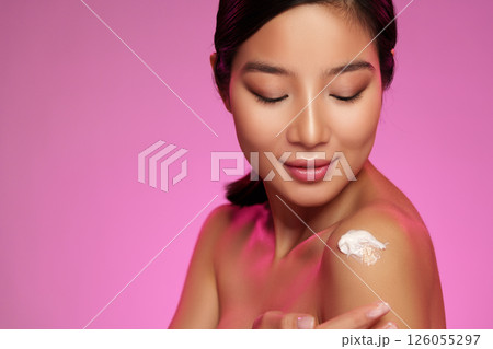 Model applying skincare product on her shoulder in a vibrant pink-themed studio setting 126055297