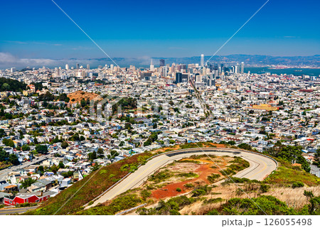 Panoramic view of downtown San Francisco from Twin Peaks, featuring iconic skyscrapers, urban density, coastal fog, and the curving summit roadway under a clear summer sky. 126055498