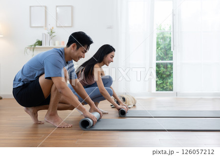 Couple rolls up their yoga mats after completing a morning workout session at home. Their pug lies nearby, enjoying the cozy atmosphere of bright living space. Couple rolls up their yoga mats after completing a morning workout session at home. Their pug lies nearby, enjoying the cozy atmosphere of bright living space. 126057212