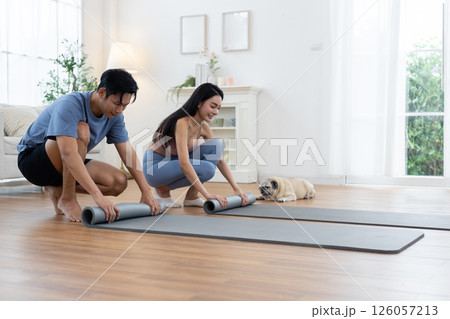 Couple rolls up their yoga mats after completing a morning workout session at home. Their pug lies nearby, enjoying the cozy atmosphere of bright living space. Couple rolls up their yoga mats after completing a morning workout session at home. Their pug lies nearby, enjoying the cozy atmosphere of bright living space. 126057213