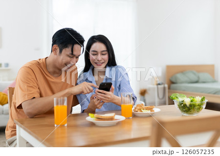 Couple enjoying together while laughing look at smartphone in dining table with glasses of orange juice and breakfast items are present in bright bedroom background. Couple enjoying together while laughing look at smartphone in dining table with glasses of orange juice and breakfast items are present in bright bedroom background. 126057235