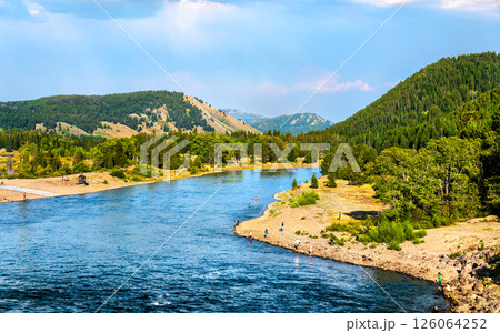 Anglers line the peaceful banks of the Snake River beneath forested hills in Grand Teton National Park in Wyoming, enjoying fly fishing on a sunny summer day with scenic mountain views. 126064252