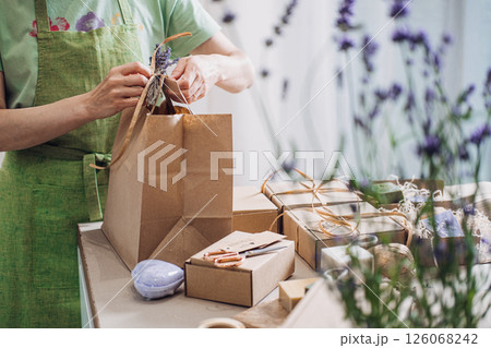 Person in green apron tying twine around a paper bag among handmade soap, scissors, and cardboard boxes. Eco-packaging, artisanal production, small business branding, sustainable consumerism Person in green apron tying twine around a paper bag among handmade soap, scissors, and cardboard boxes. Eco-packaging, artisanal production, small business branding, sustainable consumerism 126068242