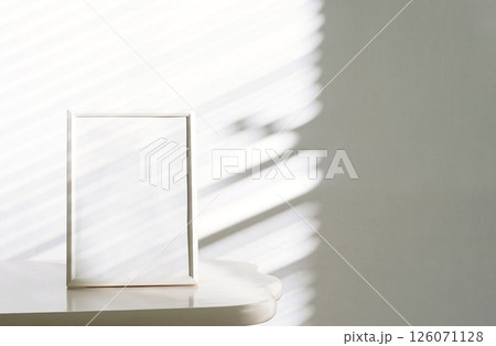 White Picture Frame Rests on Table, Natural Sunlight that Casts Gentle Shadows Through Window Blinds. White Picture Frame Rests on Table, Natural Sunlight that Casts Gentle Shadows Through Window Blinds. 126071128
