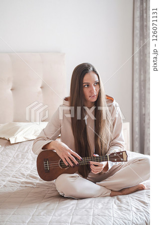 Woman with Long Hair Sits Cross Legged on Bed, Strumming a Ukulele, Enjoying Her Musical Moment. Woman with Long Hair Sits Cross Legged on Bed, Strumming a Ukulele, Enjoying Her Musical Moment. 126071131
