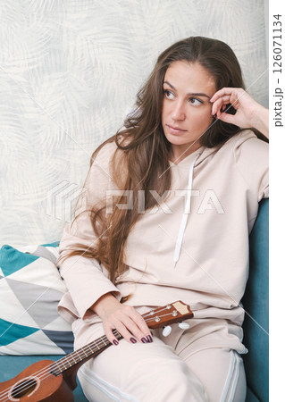 Woman with Long Hair Sits Thoughtfully on Couch, Holding Ukulele. She Gazes into the Distance, Creating Relaxed Atmosphere. Woman with Long Hair Sits Thoughtfully on Couch, Holding Ukulele. She Gazes into the Distance, Creating Relaxed Atmosphere. 126071134