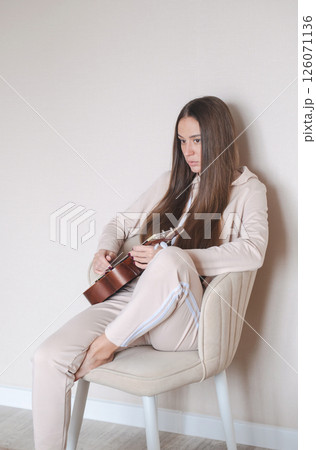 Woman Sits Comfortably on a Chair, Playing Ukulele Quietly. She Wears Casual Clothes and Seems Lost in Her Music. Woman Sits Comfortably on a Chair, Playing Ukulele Quietly. She Wears Casual Clothes and Seems Lost in Her Music. 126071136