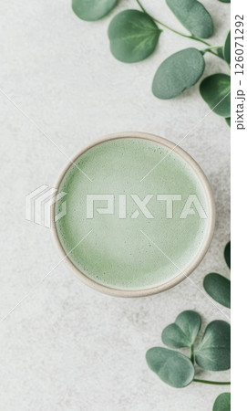 A cup of frothy green matcha latte sits on a light grey surface, adorned with fresh eucalyptus leaves 126071292