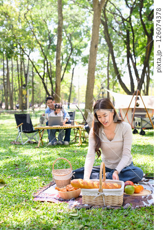Woman arranging food for a picnic while her family enjoys time at a forest campsite Woman arranging food for a picnic while her family enjoys time at a forest campsite 126074378