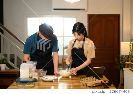 Happy couple in aprons following a recipe while making pancakes at home 126074408