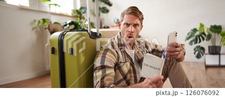 Man looks very confused, using smartphone, holding passport, sitting near luggage, planning his journey or holiday Man looks very confused, using smartphone, holding passport, sitting near luggage, planning his journey or holiday 126076092