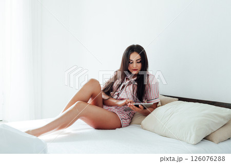 Beautiful brunette woman in pajamas with internet tablet in white bedroom on bed sleep 126076288