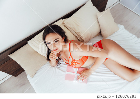 Brunette woman in red lingerie with pink gift in bedroom on bed in morning 126076293