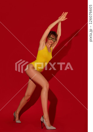 Fit woman with long legs in yellow swimsuit extends arms above head in motion wearing green sunglasses and heels against red studio background Fit woman with long legs in yellow swimsuit extends arms above head in motion wearing green sunglasses and heels against red studio background 126076605