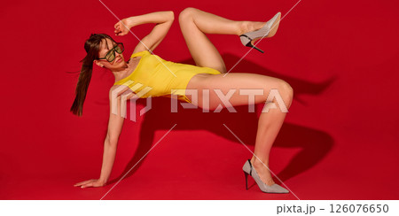 Beautiful woman with athletic body performing high leg motion on hand balance in striped heels and yellow swimsuit against red studio background. Beautiful woman with athletic body performing high leg motion on hand balance in striped heels and yellow swimsuit against red studio background. 126076650