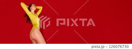 Side view of woman stretching arms behind head in yellow swimsuit showing silhouette posture against red background with negative space. 126076730