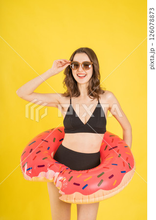 Stylish young woman in black bikini holding pink donut pool float and wearing sunglasses on yellow background, smiling and posing playfully for summer travel, swimwear, or vacation-themed concepts Stylish young woman in black bikini holding pink donut pool float and wearing sunglasses on yellow background, smiling and posing playfully for summer travel, swimwear, or vacation-themed concepts 126078873