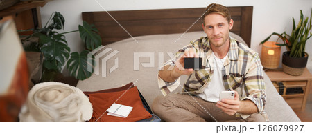 Portrait of tourist, man packing suitcase with clothes and passport, holding mobile phone, showing his credit card 126079927