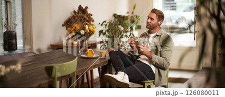 Portrait of young handsome man in casual clothes, sits in cafe and enjoys favourite song, listens to music in wireless headphones, vibing to the relaxing tunes in earphones, holding smartphone Portrait of young handsome man in casual clothes, sits in cafe and enjoys favourite song, listens to music in wireless headphones, vibing to the relaxing tunes in earphones, holding smartphone 126080011