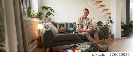 Happy man with pen and notebook, checking list of items and packing suitcase for journey, going on holiday, preparing for summer vacation, sitting in living room Happy man with pen and notebook, checking list of items and packing suitcase for journey, going on holiday, preparing for summer vacation, sitting in living room 126080020
