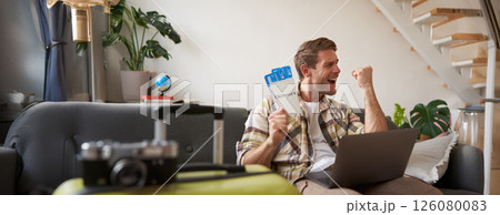 Upbeat man, tourist with two tickets, makes a fist pump, has laptop opened, goes on vacation, excited about holiday Upbeat man, tourist with two tickets, makes a fist pump, has laptop opened, goes on vacation, excited about holiday 126080083