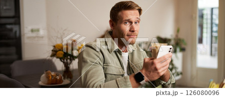 Portrait of young man in cafe, holding cup of coffee and mobile phone, looking at camera with skeptical face and a smirk 126080096