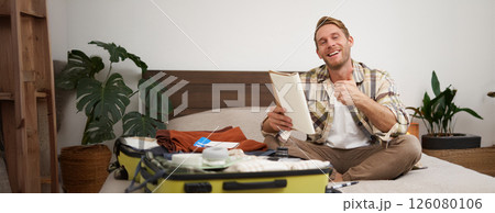 Cheerful young man, tourist with check list, showing his notes in notebook, thumbs up, packing suitcase, going on holiday, travelling abroad and preparing luggage 126080106