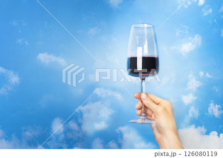 A woman's hand holds a glass of red wine against a blue sky with clouds, copy space banner A woman's hand holds a glass of red wine against a blue sky with clouds, copy space banner 126080119