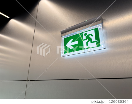 Glowing green emergency exit sign with arrow and running person symbol on brushed metal wall. Evacuation indicator in modern public building. Concept of alert, escape route and safety compliance 126080364