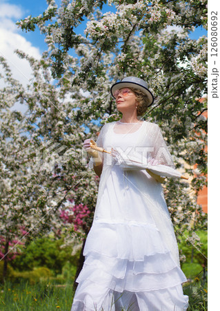 Mature woman of 60 years old in 18th century style white canotier hat, retro white dress, gloves, glasses and umbrella in blossoming white and pink apple orchard on summer day 126080692