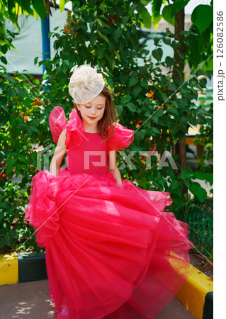 A girl in a lush pink dress and hat near the green bushes 126082586