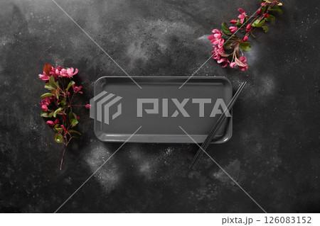 Minimalist scene of empty rectangular plate for asian food with black chopsticks on dark textured surface with spring pink flowers. Asian style. View from above. Copy space. Japanese food concept. 126083152
