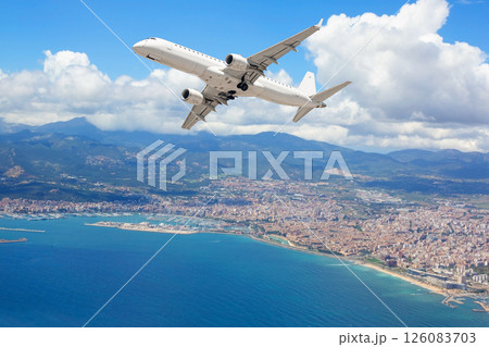 White passenger airplane flying over coastal city, mountains and blue sea White passenger airplane flying over coastal city, mountains and blue sea 126083703