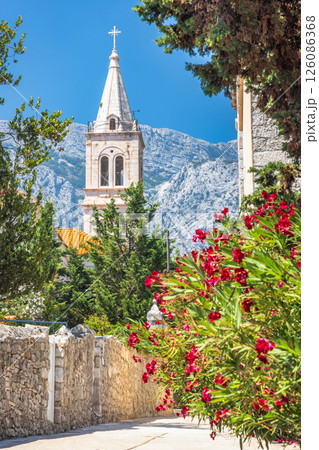 Orebic town at coast of the Peljesac peninsula in Croatia, Europe. Picturesque European church tower framed by lush foliage and a striking mountain backdrop under a clear blue sky. 126086368