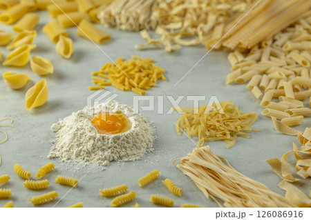 Variety of uncooked pasta with flour and egg yolk forming a circle on a gray background Variety of uncooked pasta with flour and egg yolk forming a circle on a gray background 126086916
